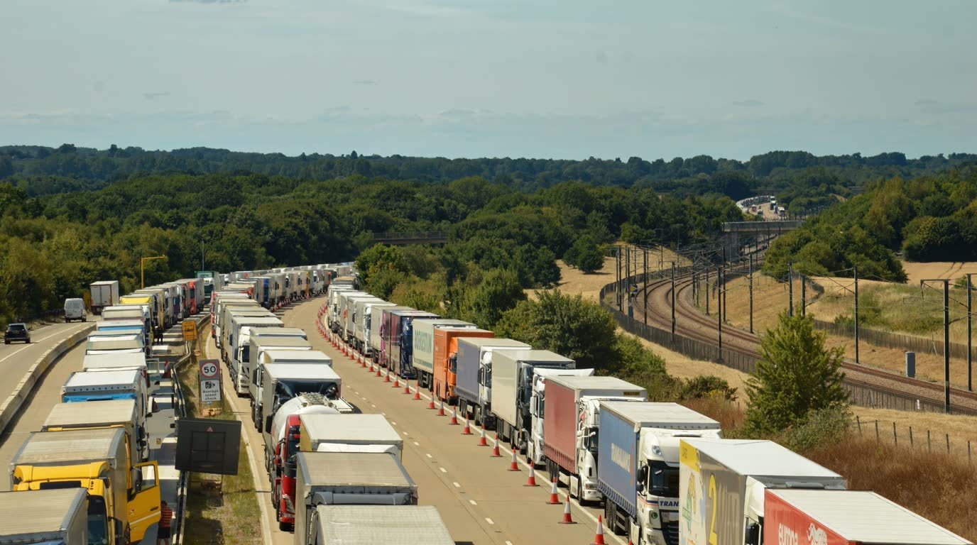 Longa fila de caminhões e carretas parados lado a lado em uma rodovia de pista dupla, separados por cones, à espera de liberação